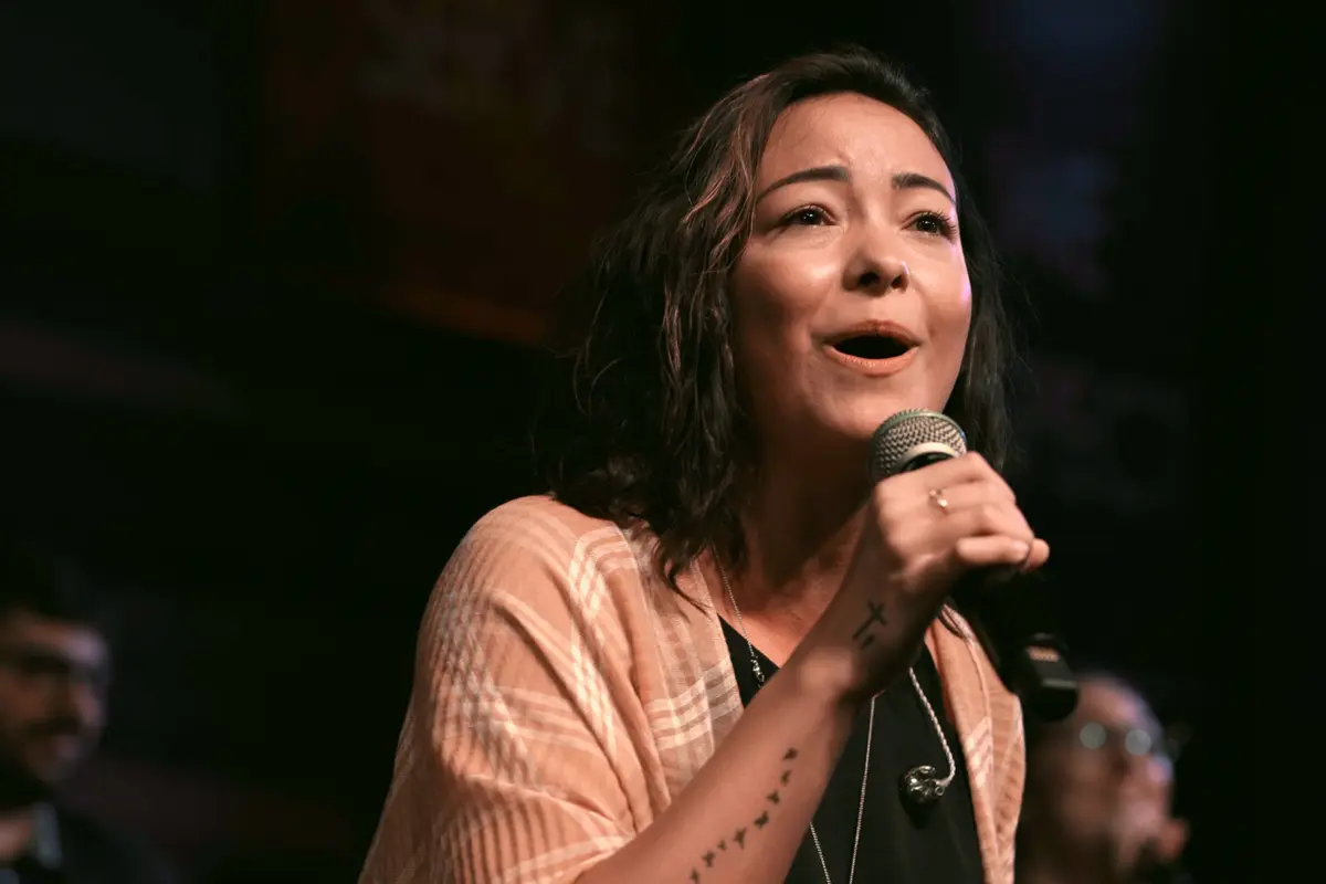 "Woman singing into a microphone on stage with a blurred background, wearing a light shawl, expressive face, indoor concert or performance setting, audience and other musicians faintly visible."